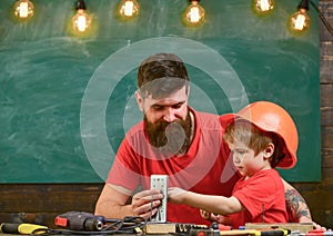 Father with beard teaching little son to use tools in classroom, chalkboard on background. Boy, child busy in protective