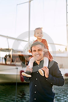 Father is carrying his baby girl on his shoulders in front of a big boat in a marina