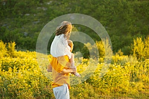 Father is carrying his daughter on shoulders walking on a field near forest in sunlight