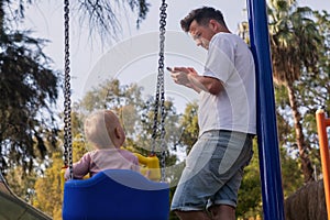 Father with child on a playground looking on phone ignoring his baby swinging.