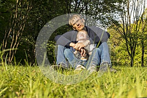 Father And Child, Son Sitting On Grass In Park, Enjoying Time Together. Summer Time