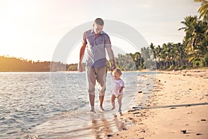 Father and child walking together on the beach coast during sunset with shining sea on background