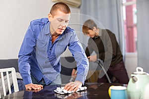 Father cleans the table with a rag while son vacuums room