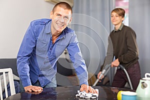 Father cleans the table with a rag while son vacuums room