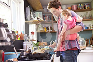 Father Cooking Meal Whilst Holding Daughter In Kitchen