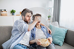 Father covering his son& x27;s eyes from inappropriate content while watching movie on tv with popcorn