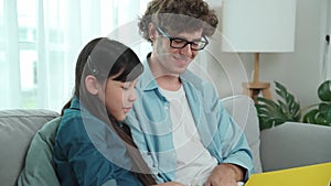 Father and cute daughter reading story together while sitting at sofa. Pedagogy.