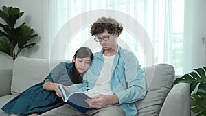 Father and cute daughter reading story together while sitting at sofa. Pedagogy.