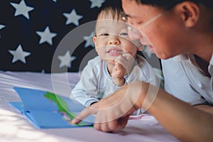 Father and Cute little Asian 2 - 3 years old toddler boy child reading bedtime story book, lying in the bed at home