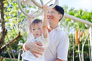 Father and Cute little Asian 2 year old toddler baby boy child having fun exercising outdoor and dad help catch up on Monkey Bars