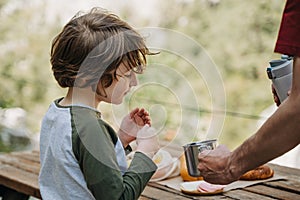 Father dad pours hot coffee tea from thermos into the mug on a family picnic in the mountains. Child school boy kid is