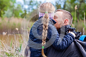 Father and daughter hugging sitting by the lake with nature lanscape. Little blond girl with braid hair walking with dad