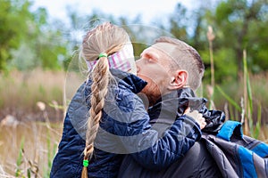 Father and daughter hugging sitting by the lake with nature lanscape. Little blond girl with braid hair walking with dad