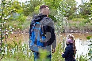 Father and daughter hugging sitting by the lake with nature lanscape. Little blond girl with braid hair walking with dad