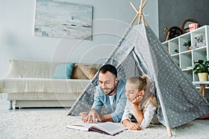 father and daughter lying on floor inside of teepee and reading book together