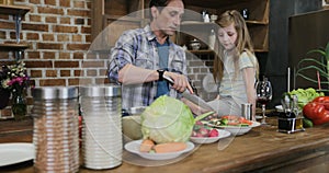 Father And Daughter Salting Vegetables Cooking Food Together While Mother And Son Use Tablet Computer Sitting At Kitchen