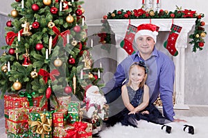 Father and daughter sit near the Christmas tree. Father have a Santa Claus hat on his head. A fireplace behind them