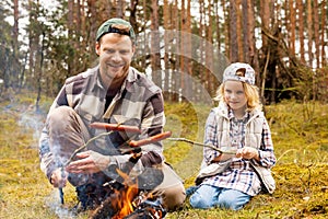 father and daughter spend time together and frying sausages over a bonfire while camping in forest