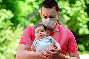 Father in facial mask holding in arms little baby daughter in summer day. Green leaves in background. Parenting during quarantine