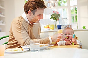 Father Feeding Baby Sitting In High Chair At Mealtime