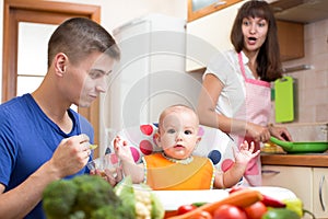 Father feeding his baby while mother cooking at kitchen