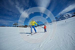 Father go downhill teach child to ski connected by poles