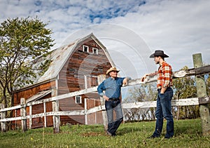 Father and grown son wearing cowboy hats standing and chatting by a red barn.