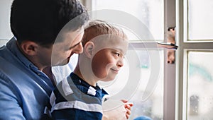 Father with happy down syndrome son indoors at home, looking through window.