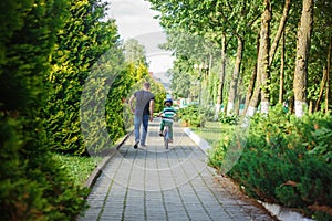 Father help son to ride bicycle in summer park. Back view