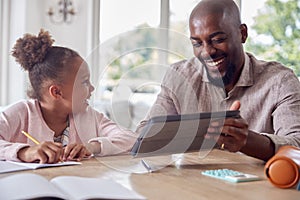 Father Helping Daughter With Home Schooling Sitting At Table With Digital Tablet