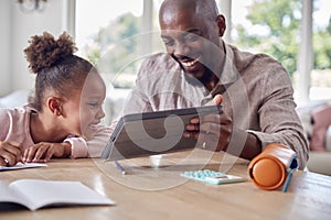 Father Helping Daughter With Home Schooling Sitting At Table With Digital Tablet