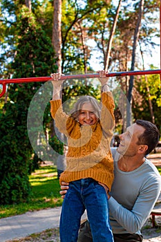 Father helping daughter to pull up on the uneven bars at the playground. Healthy family concept