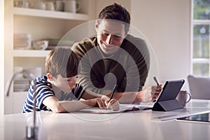 Father Helping Son With Homework Sitting At Kitchen Counter Using Digital Tablet Together