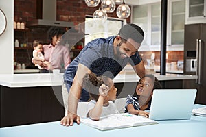 Father Helps Children With Homework Whilst Mother With Baby Uses Laptop In Kitchen