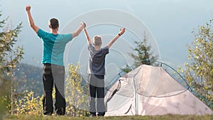 Father and his child son walking near camping tent with raised hands while hiking together in summer mountains. Active family trav