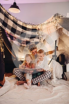 Father and his kids reading book together while sitting in handmade tent in children's room