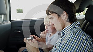 Father with his son sitting in car seat and playing on smart phone while having car ride in backseat