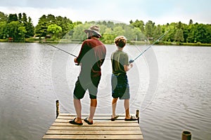 Father and his young son are on a dock on a small lake fishing while on summer vacation.