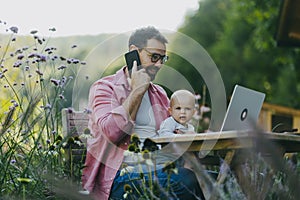 Father holding baby while working on laptop outdoors, in garden. Businessman working remotely from homeoffice, making