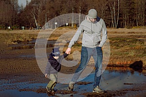 Father holding his little son`s hand walking alone beach on wet sand. Sun and grass reflecting in piddles.