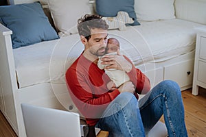 Father holding his newborn crying baby during working on laptop.