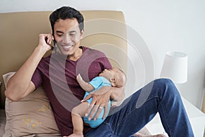 father holding his newborn sweet adorable baby sleeping on his arms while using mobile phone on the bed