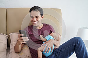 father holding his newborn sweet adorable baby sleeping on his arms while using mobile phone on the bed