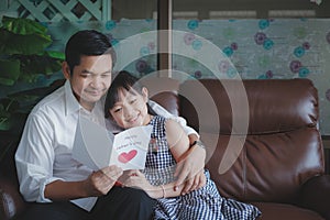 Father hugging daughter and reading the postcard that his daughter happily gave inside the room.Concept of happy father`s day