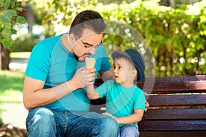 Father and little son eating ice-cream together in park in sunny summer or autumn day. Boy offers ice-cream to daddy. Happy family