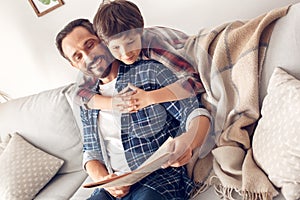 Father and little son at home boy hugging dad sitting on sofa reading newspaper cheerful