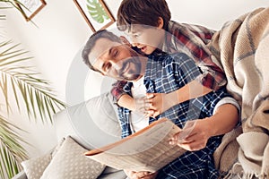 Father and little son at home boy hugging surprised dad sitting on sofa with newspaper looking at son playful