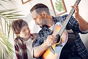 Father and little son at home sitting on sofa boy smiling looking at dad playing guitar