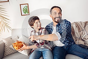 Father and little son at home sitting on sofa boy taking away potato chip from dad playful watching tv