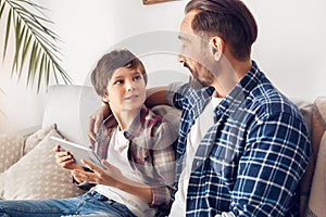 Father and little son at home sitting on sofa dad hugging boy with digital tablet talking joyful